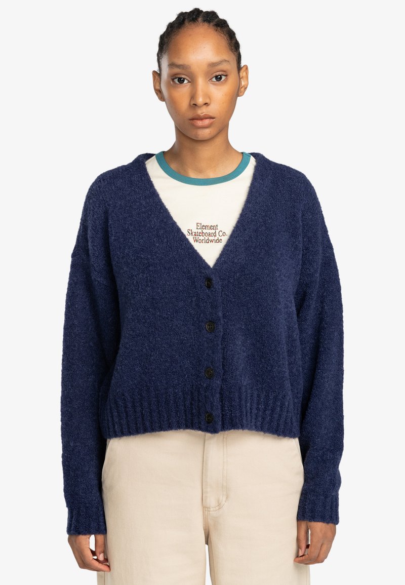 Marineblauwe fluffy cardigan met V-hals, lange mouwen en vijf zwarte knopen. Geribbelde zoom en boorden, gedragen over een crèmekleurig overhemd.