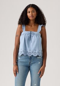 Light blue sleeveless top with ruffled straps, tie-front detail, and scalloped lace hem featuring floral cutouts. Paired with distressed jeans.