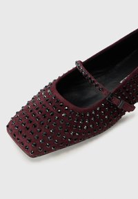 Chaussure plate en daim bordeaux avec un bout carré, ornée d'un motif clouté noir et d'une sangle décorative avec une boucle.