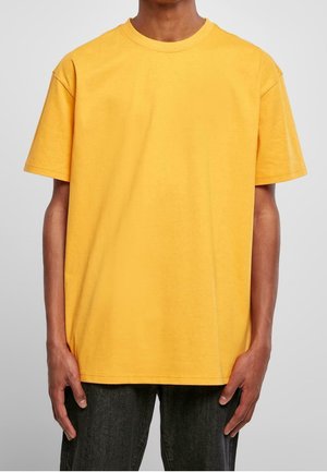 Personne portant un t-shirt jaune uni à manches courtes et un pantalon foncé, debout les bras détendus le long du corps sur un fond gris.