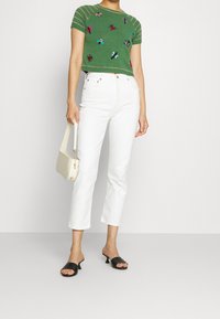 Suéter de punto verde de manga corta con bordado floral multicolor, combinado con jeans blancos cortos y zapatos negros tipo slip-on. Bolso de hombro color crema.