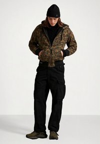 Casaco com padrão camuflado com fecho de correr e capuz, malha preta por baixo, calças cargo pretas e sapatos verde-oliva. Tecido texturizado, ajuste casual.