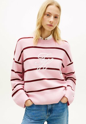 Lyshåret kvinde iført en pink sweater med vandrette burgunderstriber og hvide broderede initialer, kombineret med blå jeans og hænderne i lommerne.