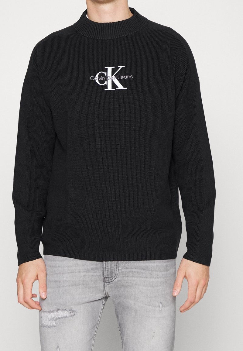 Homme portant un pull noir Calvin Klein Jeans avec un logo blanc et un jean gris clair déchiré sur un fond uni.