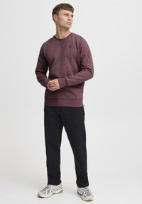 Bordeaux sweatshirt med tekstureret stof, rund halsudskæring og et lille logo på brystet. Bæret med mørke bukser og grå sneakers.