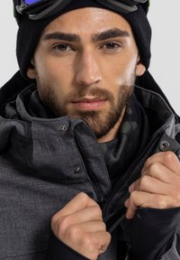 Men's black textured jacket with a zippered front, fastened collar, and patterned inner lining. A beanie and goggles complete the outfit.