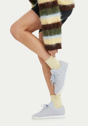 Person iført lysegule sokker og grå sneakers, med det ene ben bøjet og hånd, der rører anklen, iført en stribet, fluffy sweater.