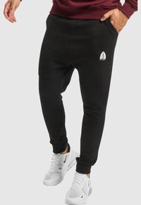 Black sweatpants featuring a soft texture, tapered design, and a white logo on the left thigh, paired with white athletic shoes.