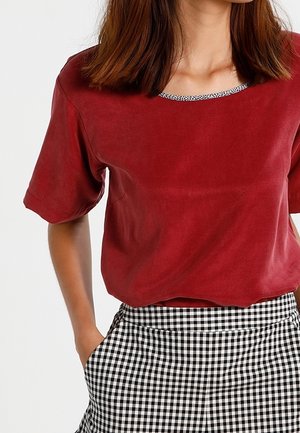 Czerwony zamszowy top z krótkim rękawem i okrągłym dekoltem z czarno-białą wzorzystą lamówką. W połączeniu z czarno-białymi spodniami w kratę.