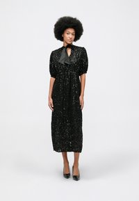 Žena s afro frizurom nosi crnu midi haljinu sa šljokicama i napučenim rukavima, uz crne visoke potpetice, stojeći na jednobojnoj pozadini.