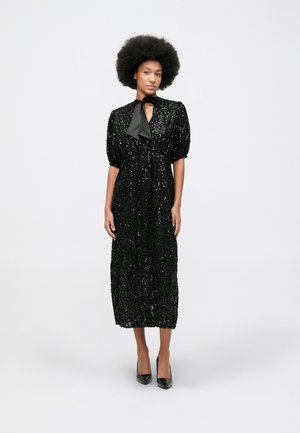 Mulher com cabelo afro, vestindo um vestido midi preto com lantejoulas, mangas bufantes e saltos altos pretos, de pé contra um fundo simples.