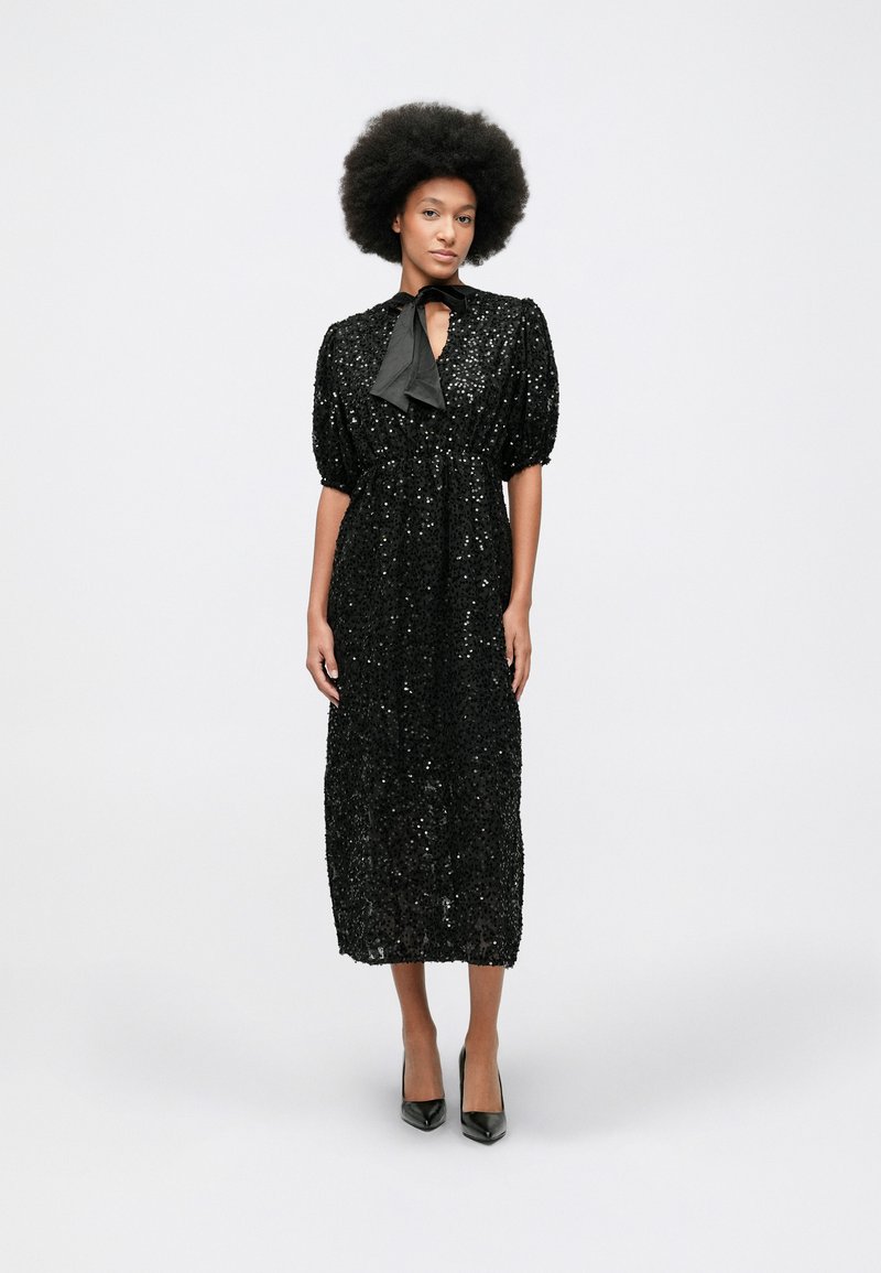 Žena s afro frizurom nosi crnu midi haljinu sa šljokicama i napučenim rukavima, uz crne visoke potpetice, stojeći na jednobojnoj pozadini.