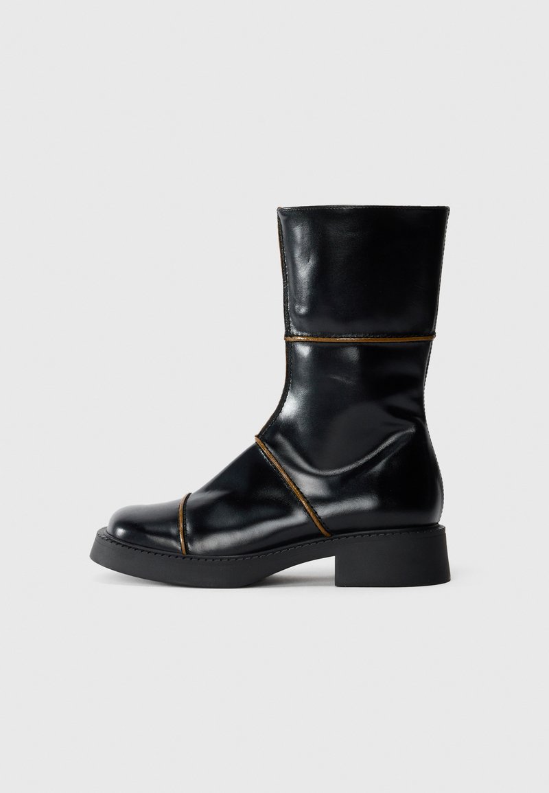 Svart läderankelboots med glansig finish, rund tå, låg blockklack och kontrasterande bruna sömnadsdetaljer. Slät textur och stilren design.