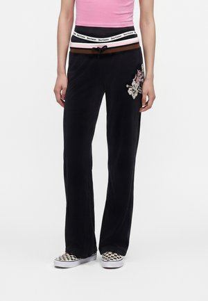Personne portant un pantalon en velours noir avec une broderie florale, un haut rose et des chaussures slip-on à carreaux, debout devant un fond blanc.