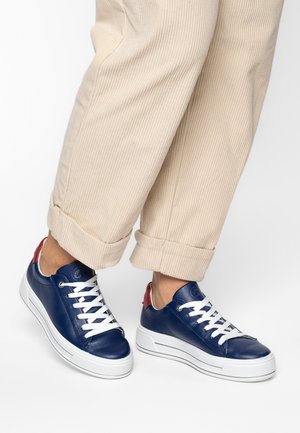 Person wearing beige rolled-up corduroy pants and navy blue sneakers with white laces and soles, and red heel accents.