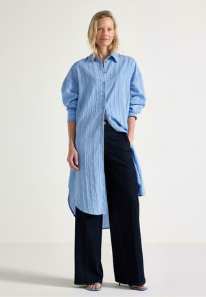 Blåstribet langærmet skjorte med knaplukning foran, med buet kant og afslappet pasform, kombineret med mørke bukser med vid ben.