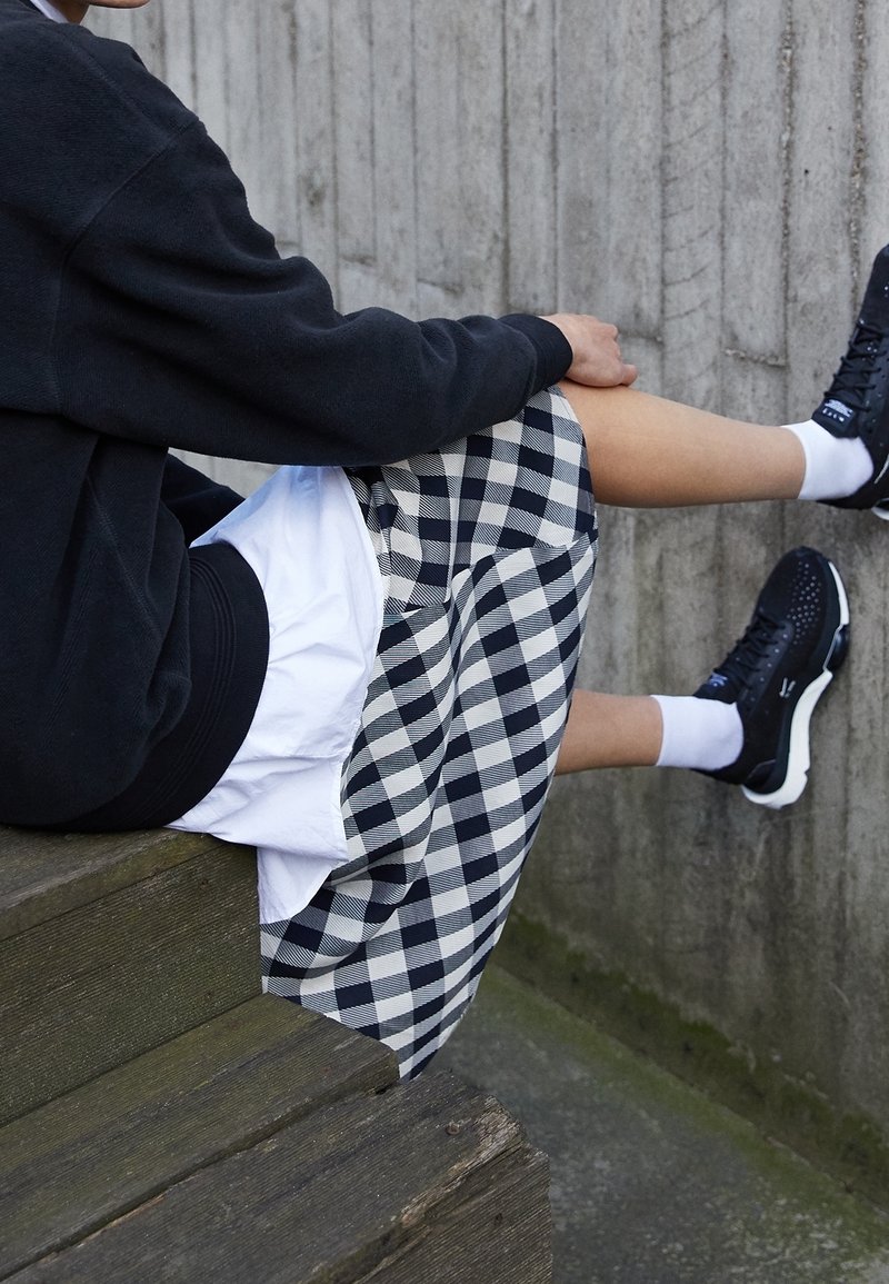 Personne assise sur des marches en bois portant un sweat-shirt noir, un short à carreaux, des chaussettes blanches et des baskets noires devant un mur en béton.