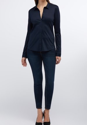 Kobieta ubrana w dopasowaną granatową koszulę zapinaną na guziki, ciemne obcisłe dżinsy i czarne szpilki, stojąca na jednolitym tle.