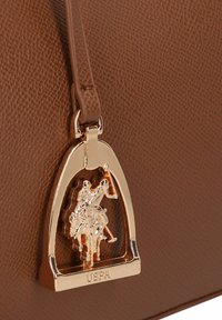 Brązowa teksturowana torebka z metaliczną złotą zawieszką w kształcie jeźdźca konnego, z logo "USPA" wkomponowanym w projekt.