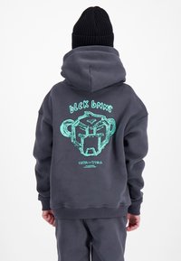 Persoon draagt een donker hoodie en broek, een zwarte beanie, met een turquoise afbeelding van een gestileerd gezicht en de tekst "Black BNNS" op de achterkant van de hoodie.