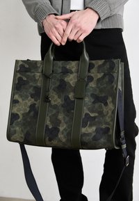 Borsa tote mimetica verde con dettagli neri, caratterizzata da un design strutturato, doppi manici superiori, tracolla staccabile e tessuto texturizzato.