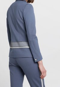 Niebieska kurtka o slim fit, z srebrnymi detalami w talii i długimi rękawami. Materiał wydaje się gładki, z dopasowanym krojem.