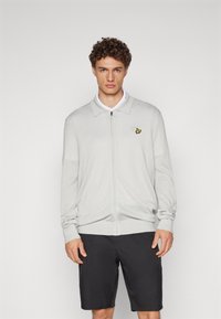 Light gray zip-up cardigan with long sleeves, ribbed cuffs, and collar; features a small yellow logo on the left chest. Worn with black shorts.