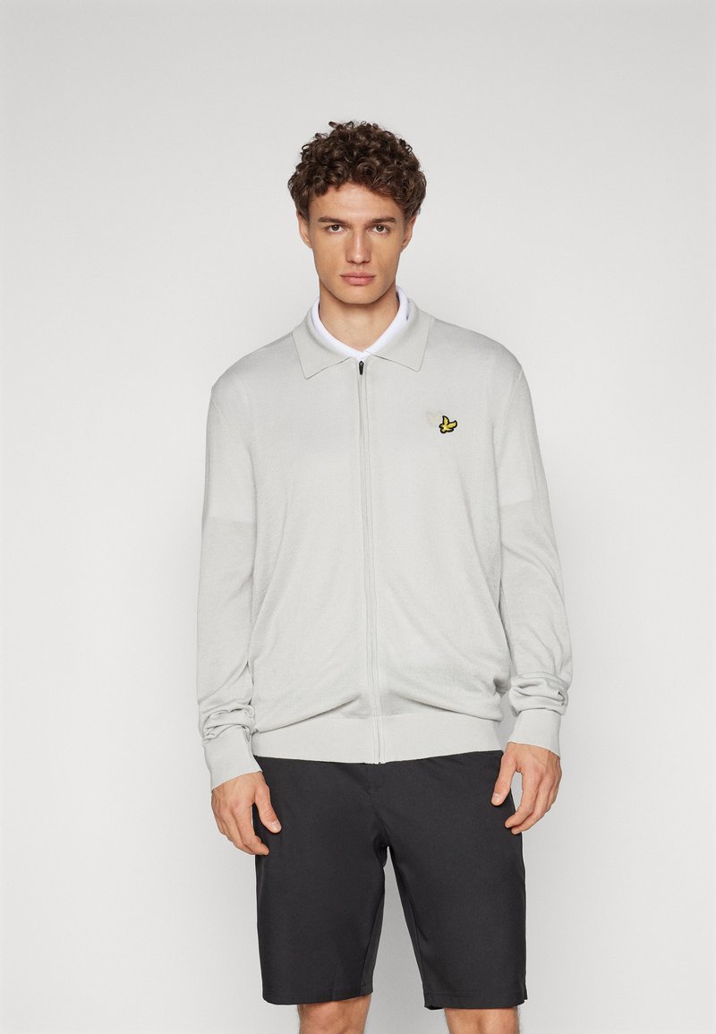 Cardigan zippé gris clair avec des manches longues, des poignets côtelés et un col ; présente un petit logo jaune sur la poitrine gauche. Porté avec un short noir.