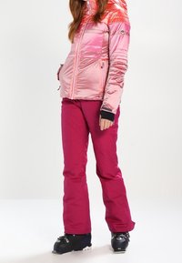 Roze donsjack met horizontale strepen, ritszakken en zwarte manchetten; gecombineerd met felroze skibroek en zwarte skischoenen.