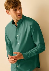 Cămașă lungă cu mâneci, de culoare teal, cu guler cu nasturi, având o căptușeală interioară cu model și o textură netedă. Modelul își gesticulează mâinile.