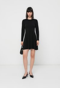 Woman in black long-sleeve dress, black heels, holding small black handbag, standing against plain white background.
