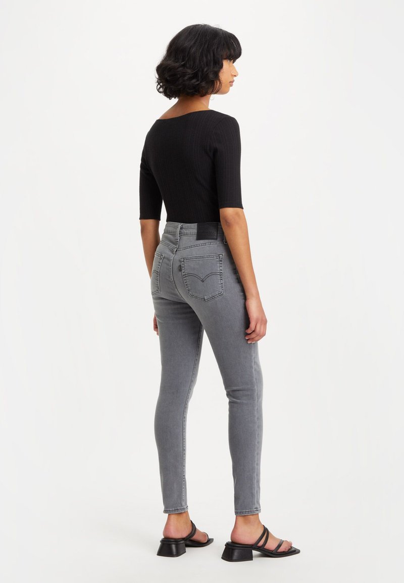 Schwarzes, tailliertes Oberteil mit gerippter Textur, kombiniert mit hochtaillierten grauen Skinny-Jeans mit Gesäßtaschen. Schwarze, hochhackige Sandalen runden den Look ab.