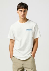 Camiseta de algodón blanca con cuello redondo y mangas cortas. Presenta un logo azul de "Wrangler" en el lado izquierdo del pecho. Corte casual.