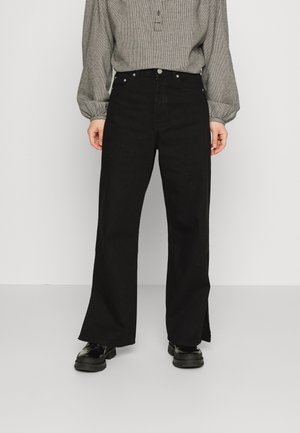 Personne portant un pantalon noir taille haute à jambes larges avec des fentes latérales, associé à un haut gris à manches longues et des chaussures noires épaisses.