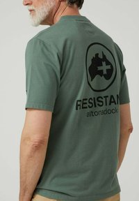 Hombre mayor con barba canosa que lleva una camiseta verde con un estampado negro del mapa de Australia y una cruz, y el texto "RESISTAN altonadock" en la parte de atrás.