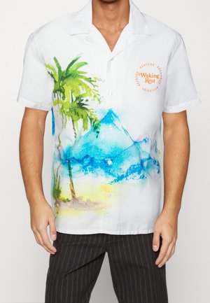 Chemise blanche à manches courtes avec boutons, imprimé aquarelle de palmier et montagne bleue, texte orange "Waking Rest" sur la poitrine, portée avec un pantalon rayé foncé.