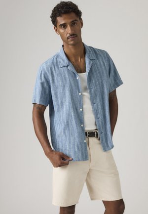 Hombre vistiendo una camisa azul de manga corta con botones sobre una camiseta blanca sin mangas y pantalones cortos beige con cinturón trenzado, posando frente a un fondo liso.