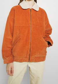 Veste en velours côtelé orange avec col en fausse fourrure, dotée d'une fermeture éclair à l'avant et de poches latérales, conçue avec une coupe décontractée et une texture côtelée.