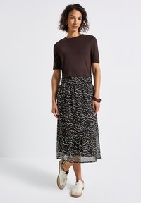 Femme se tenant debout portant un haut à manches courtes marron foncé, une jupe midi à motifs noir et blanc, des baskets blanches et des bracelets au poignet.