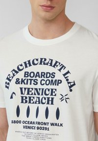 Weißes Baumwoll-T-Shirt mit auffälligem navyblauen Text, der "BEACHCRAFT L.A.", "VENICE BEACH" und grafische Elemente zeigt. Rundhalsausschnitt, lässige Passform.