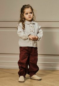 Camicia bianca con pois neri e spalle arricciate, bottoni marroni; pantaloni bordeaux con polsini elastici; scarpe chiare con punta rotonda.