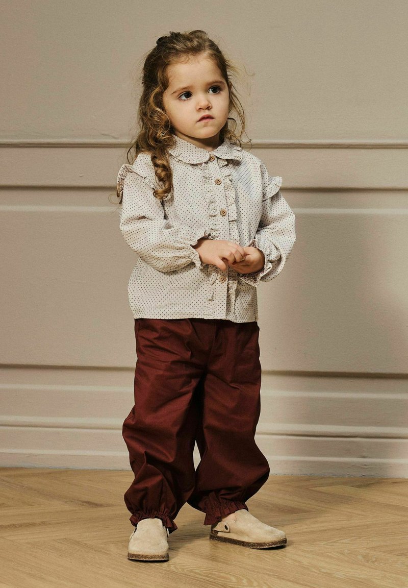 Camicia bianca con pois neri e spalle arricciate, bottoni marroni; pantaloni bordeaux con polsini elastici; scarpe chiare con punta rotonda.