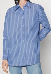 Hellblaues langärmliges Hemd mit weißen vertikalen Streifen, Brusttasche und klassischem Kragen. Hergestellt aus glattem Stoff.