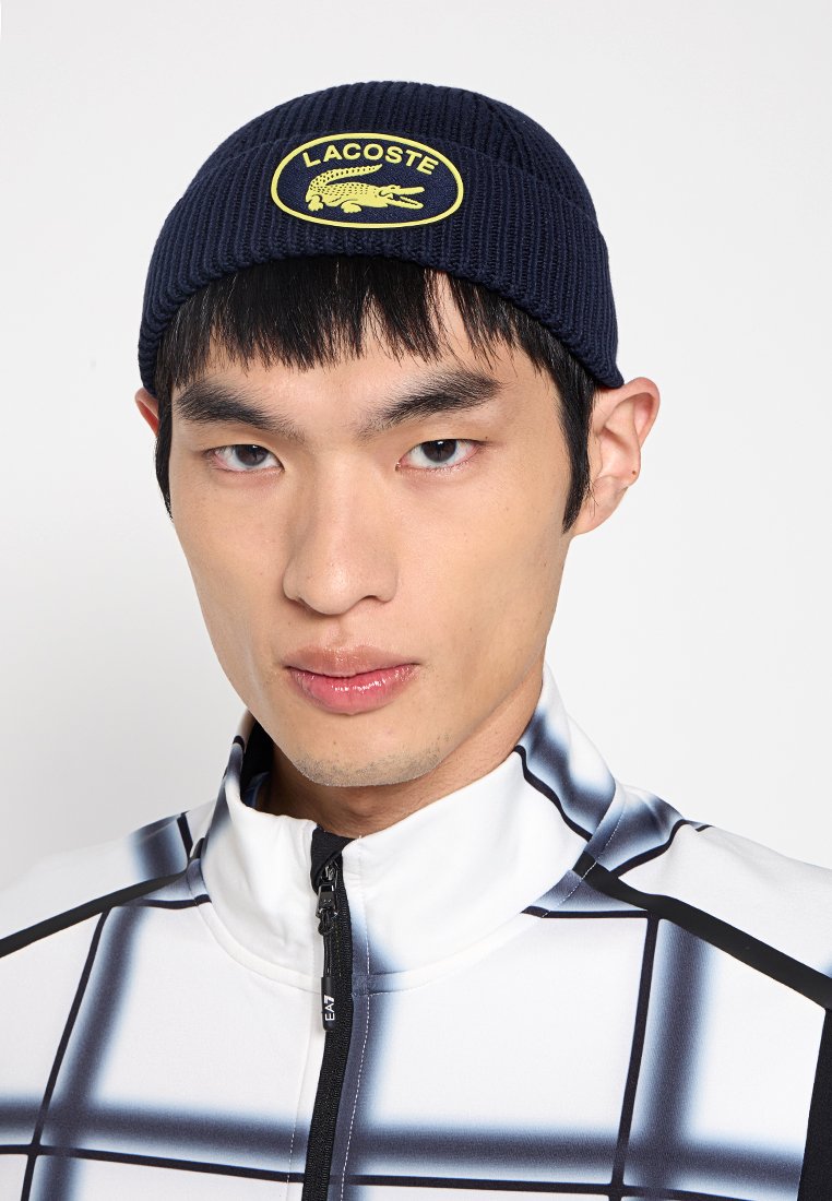 Marineblaue Rippmütze mit einem gelben Lacoste-Logo-Patch, getragen von einem Modell in einer weiß und schwarz karierten Jacke mit Reißverschluss.