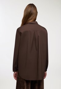 Persona con capelli lunghi e castani di spalle, che indossa una camicia larga a maniche lunghe di colore marrone scuro e pantaloni marrone scuro su uno sfondo neutro.