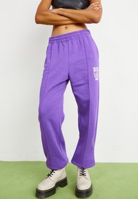 Fialové tepláky s elastickým pasem, bočními pruhy a logovou nášivkou; doplněné krémovými platformovými botami a koženým crop topem.