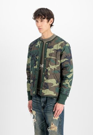Giovane uomo con una giacca trapuntata mimetica sopra una maglietta verde e jeans strappati blu, in piedi davanti a uno sfondo bianco.