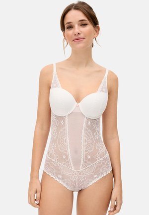 Mujer joven vestida con un body blanco de encaje con copas acolchadas, de pie contra un fondo liso y claro, mirando al frente con una sonrisa sutil.