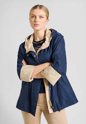 Donna con le braccia incrociate che indossa una giacca blu navy con fodera beige e dettagli sui polsini, pantaloni beige e una camicia a righe blu navy e beige.