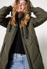 Casaco acolchoado verde-oliva com capuz, apresentando fecho de correr e tiras pretas, combinado com um suéter preto e jeans azul claro.